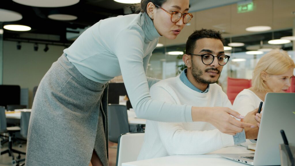 A woman is standing beside a man sitting at a computer explaining AI in the hiring process for candidates.
